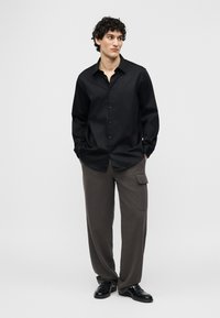 Άνδρας με σγουρά μαλλιά στέκεται με τα χέρια στις τσέπες φορώντας μαύρο πουκάμισο με κουμπιά, σκουρόχρωμο γκρι παντελόνι cargo και μαύρα επίσημα παπούτσια.