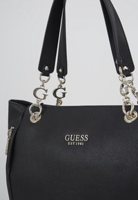 Zwarte gestructureerde Guess-handtas met zilverkleurige hardware, metalen "G"-bedels, dubbele handvatten en het "GUESS EST 1981" logo aan de voorkant.