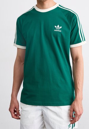 Mężczyzna w zielonej koszulce Adidas z białymi paskami na rękawach i białym wykończeniem, w połączeniu z białymi spodenkami Adidas z zielonymi paskami.