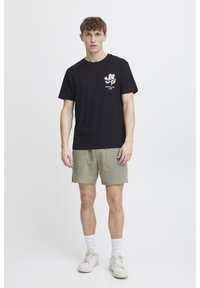 Sort bomulds t-shirt med hvid blomstret grafik på venstre bryst, parret med lysegrønne shorts. Modellen har hvide sneakers og ankelsokker på.