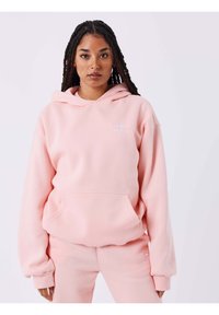 Lichtroze hoodie van zacht materiaal, voorzien van een voorzak en een klein geborduurd logo op de borst. Losse pasvorm met brede mouwen.