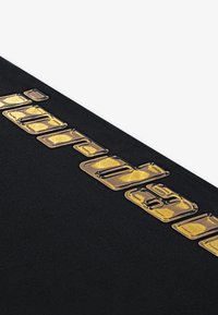Černá textilie s texturovaným, vyraženým zlatým nápisem. Písmena jsou tučná a úhlová, což vytváří výrazný kontrast s pozadím.