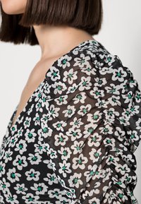 Schwarze Bluse mit floralem Muster, die weiße und grüne Blumen zeigt, aus durchsichtigem Stoff, mit gerafften Ärmeln und einem tiefen V-Ausschnitt.