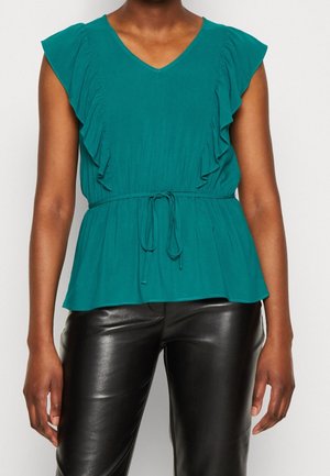 Femme portant un chemisier turquoise à manches à volants avec une taille à cordon et un pantalon en cuir noir, debout devant un fond clair uni.