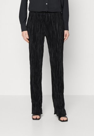 Woman wearing black pleated straight-leg pants with a black long-sleeve shirt and black open-toe square heel sandals.