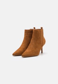 Botas de ante marrón hasta el tobillo con punta afilada, tacón delgado y paneles laterales elásticos. Textura suave con un patrón sutil.