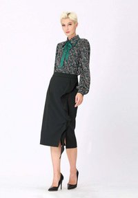 Blouse verte à motif avec nœud, associée à une jupe crayon noire avec un design asymétrique à volants et une fente latérale, le tout agrémenté de talons noirs.