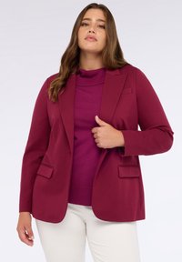 Burgundský blazer se strukturovanými klopami, dvěma předními kapsami a hladkou texturou, položený na fialový rolák a spojený s bílými kalhotami.