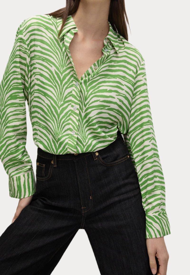 Donna che indossa una camicia button-up a strisce zebra verde e bianca, infilata in un paio di jeans scuri a vita alta.