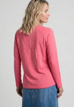 Frau mit schulterlangem blondem Haar, die ein pinkes langärmeliges Shirt mit dem Text "WE ARE ALIFE" und einen blauen Jeansrock trägt, seitlich stehend.