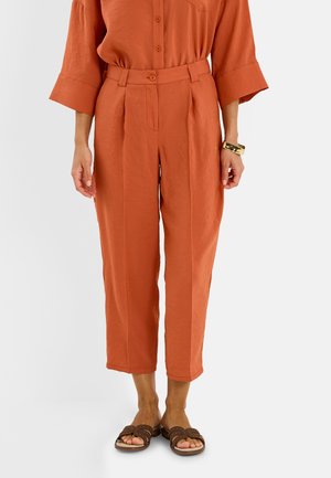 Pantalones cortos de talle alto en naranja óxido con pliegues, botón y trabillas para cinturón, usados con camisa a juego y sandalias marrones con tachuelas.