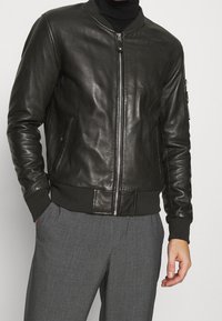 Schwarze Leder-Bomberjacke mit durchgehendem Reißverschluss, gerippte Bündchen und Saum sowie Seitentaschen. Verfügt über eine Reißverschlusstasche am linken Ärmel.