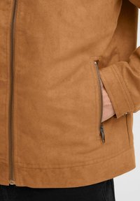 Veste en daim marron avec une fermeture éclair à l'avant et une poche latérale. Le tissu a une texture douce avec une finition mate et des détails de couture soignés.