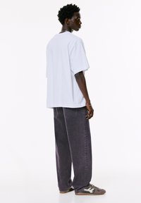 Weißen, übergroßen T-Shirt mit kurzen Ärmeln, kombiniert mit dunkelvioletten, weiten Jeans und grauen Sneakers mit cremefarbenen Akzenten. Hinteransicht, minimalistisch gestaltet.