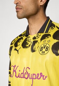 Keltainen urheilupaita, jossa on mustaa leopardikuviota, musta kaulus, vaaleanpunainen "KidSuper" -logo ja pyöreä BVB 09 -tunnus kahdella tähdellä.