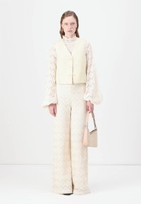Cream textured wide-leg pants with a zigzag pattern, paired with a light yellow vest and a beige handbag. Blouse has puffed sleeves.