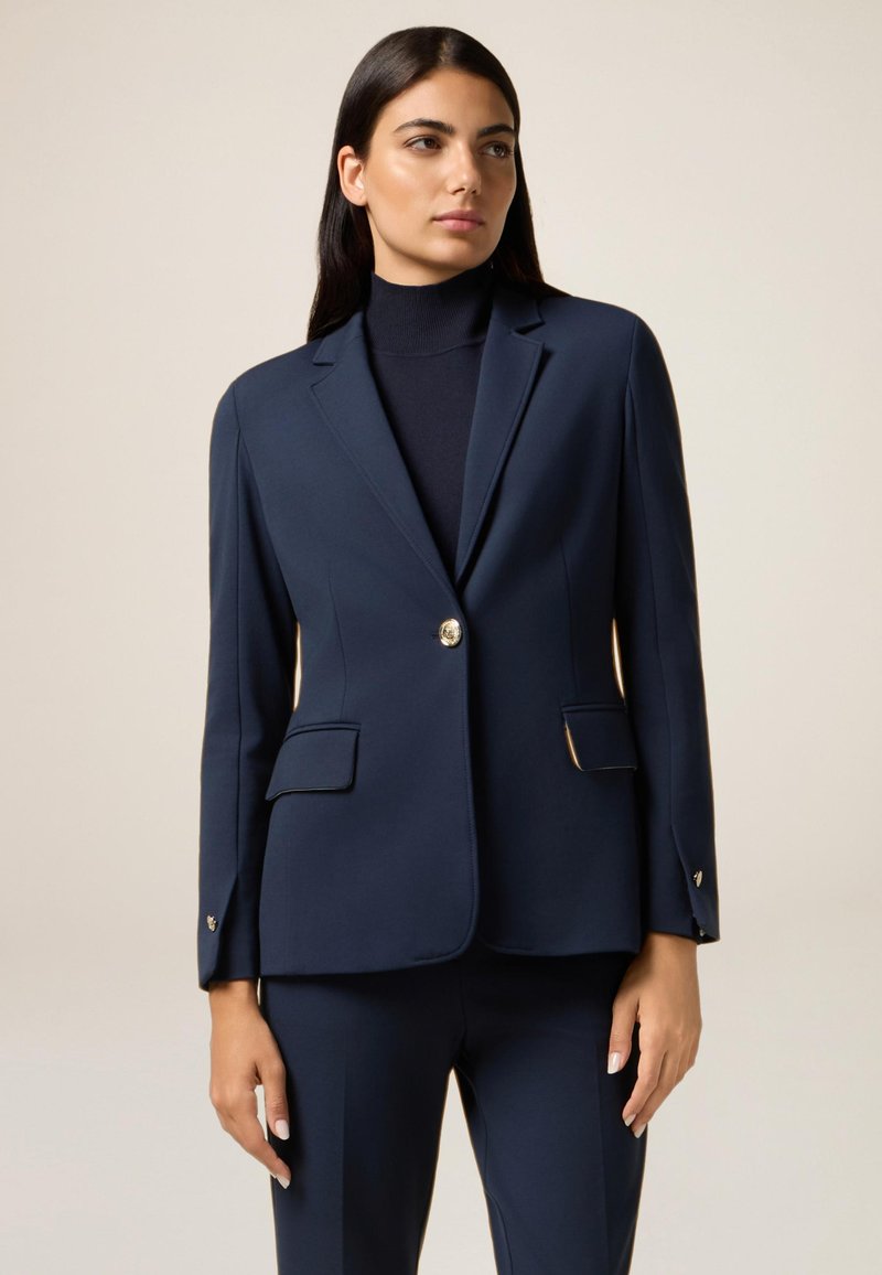Marineblå blazer i en skræddersyet design med en enkelt guldknap, strukturerede skuldre og to forlommer. Glat stof med let glans.