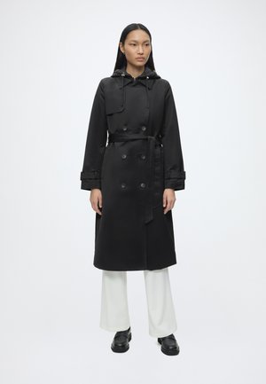 Femme portant un trench-coat noir à double boutonnage avec ceinture, pantalon blanc à jambes larges et mocassins noirs, debout devant un fond blanc uni.