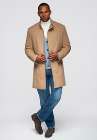 Beige Mantel mit hohem Kragen, über einem hellblauen Pullover getragen. Dunkle Jeans und braune Wildlederstiefel runden das Outfit ab. Vor einem schlichten Hintergrund.
