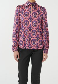 Femme portant un chemisier à motifs géométriques violet, rose et orange avec une petite encolure en forme de trou de serrure et un pantalon noir.