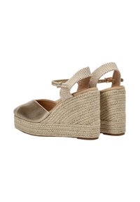 Las sandalias de cuña presentan una puntera metálica dorada y un tacón de yute trenzado, con una correa para el tobillo a rayas en color crema y un cierre. Suela plana.