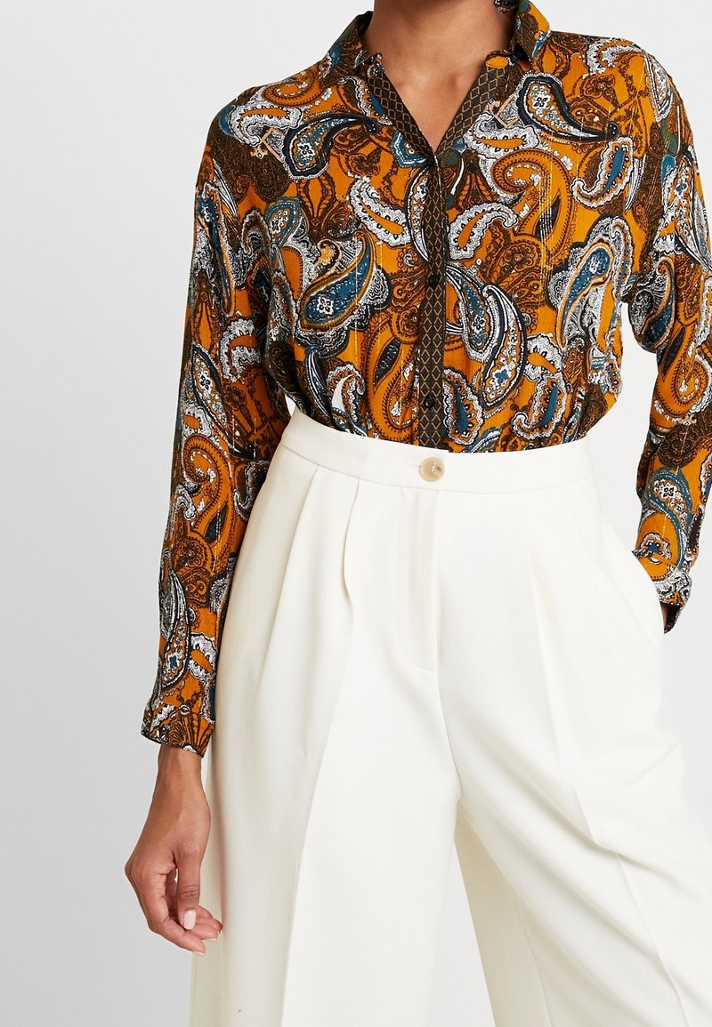 Modella che indossa una blusa a fantasia arancione e blu con colletto, abbinata a pantaloni bianchi a vita alta, mentre assume una posa.