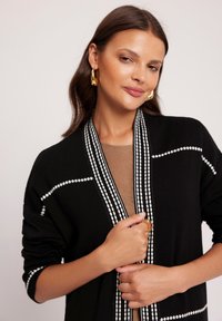 Czarny kardigan z białymi geometralnymi paskami, otwarty z przodu, o miękkiej fakturze. Noszony na beżowej bluzce, z dodatkiem złotego pierścionka.
