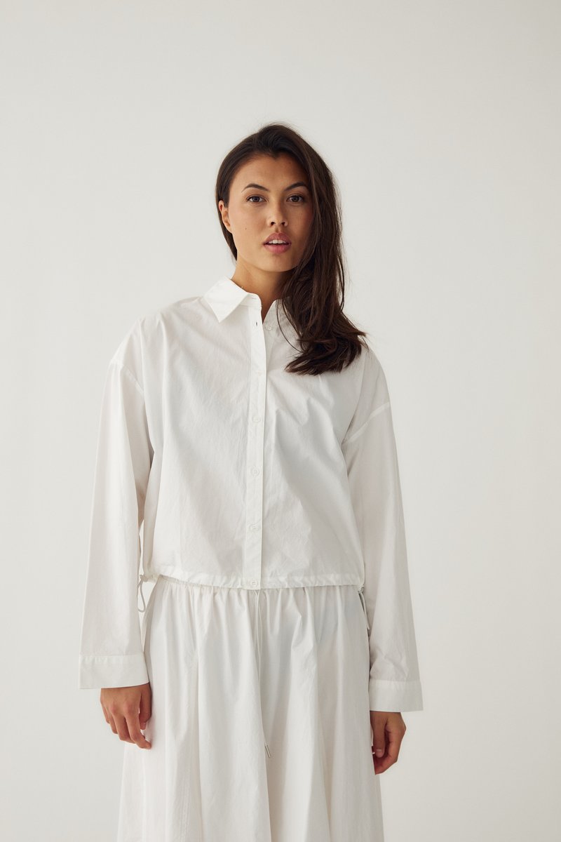 Femme aux longs cheveux foncés portant une chemise blanche ample à boutons et une jupe blanche assortie sur un fond clair uni.