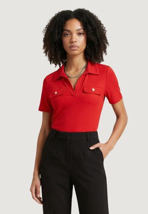 Femme aux cheveux bouclés portant un haut rouge à manches courtes avec col et poches à boutons, ainsi qu'un pantalon noir taille haute, debout avec une main dans la poche.