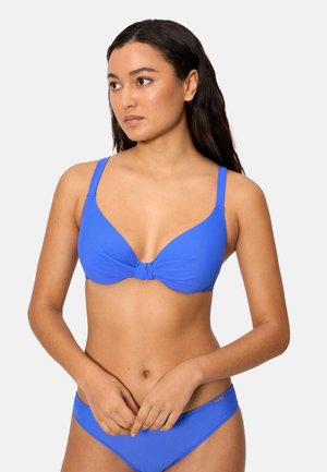 Mujer con cabello largo y oscuro, vistiendo un bikini azul brillante con un top anudado, de pie frente a un fondo liso de color gris claro.