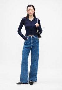 Jūras spēku zilas ribu adījuma īsās veste ar kapuci, savienota ar plašām zilām džinsu biksēm. Modelis tur melnu rokassomu, valkājot melnas kurpes.