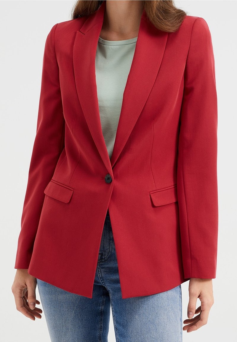 Femme portant un blazer rouge ajusté avec un bouton noir sur un haut gris clair et un jean bleu, debout devant un fond uni.