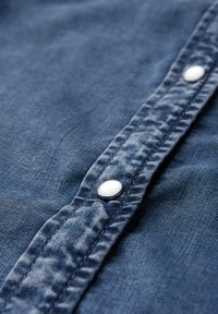Jeanshemd aus strukturiertem blauen Stoff, mit einer Reihe von metallisch silbernen Knöpfen entlang der Blende und einer genähten Nahtdetail.