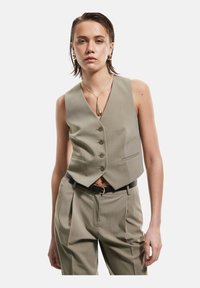 Gilet color khaki con un design sartorial, cinque bottoni e due tasche laterali. Abbinato a pantaloni a vita alta e una cintura nera.