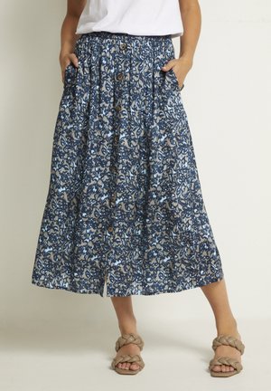 Vrouw draagt een midi rok met knopen aan de voorkant in blauw en beige met bloemmotief, zijzakken en bruine sandalen met gevlochten bandjes.