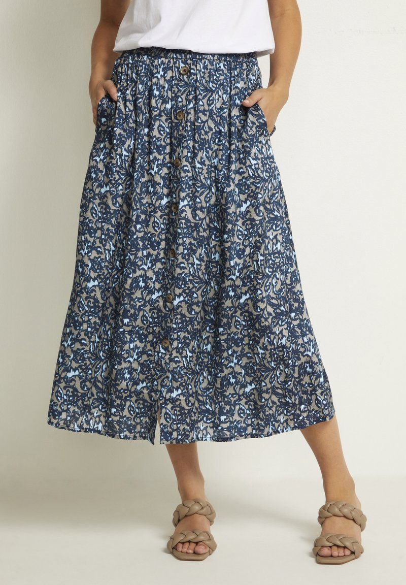 Femme portant une jupe midi boutonnée devant, à motif floral bleu et beige, avec des poches latérales et des sandales marron à brides tressées.