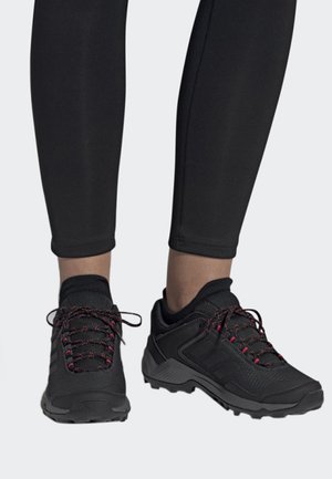 Zwarte sportieve schoenen met een geweven bovenkant van textuur, roze-rode veters en robuuste zolen, gedragen met zwarte strakke leggings.