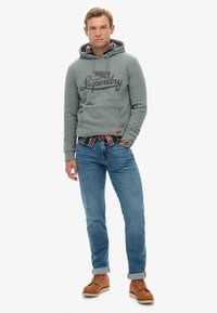 Grijze hoodie met trekkoord en logo, gedragen over een geruit overhemd, gecombineerd met een blauwe spijkerbroek en bruine veterlaarzen. Casual, comfortabele pasvorm.