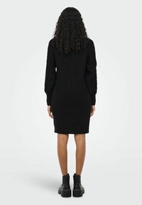 Robe noire en maille côtelée avec manches longues, silhouette ajustée et longueur mi-cuisse. Associée à des bottines noires à plateforme.