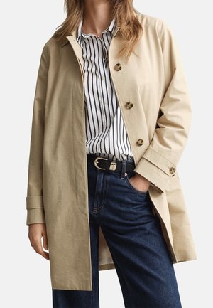 Femme portant un manteau beige boutonné par-dessus une chemise blanche à rayures verticales noires, un jean bleu foncé et une ceinture noire avec une boucle argentée.