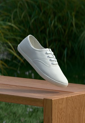 Hvide kanvas sneakers med rund tå, seks snørrehuller og tekstureret gummisål, hængende over en træoverflade med grønne græs i baggrunden.