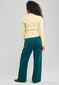 Maglietta a maniche lunghe gialla a coste abbinata a pantaloni ampi color teal con strisce bianche laterali. L'outfit presenta una texture liscia e una silhouette aderente.