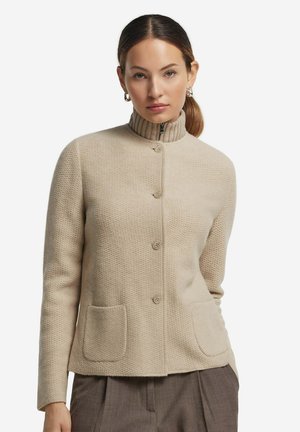Beige stickad tröja med hög ribbad krage, knappstängning fram och två framfickor. Strukturerad tyg med en figurnära siluett.