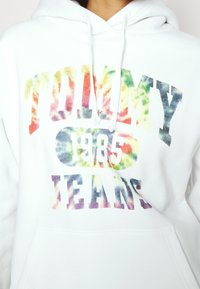 Sudadera blanca con un gráfico de tie-dye colorido que dice "TOMMY JEANS 1985" en letras grandes. Incluye un bolsillo frontal y capucha con cordón.