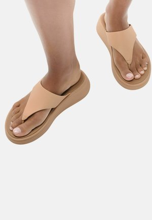 Voeten met beige teenslippers met dikke zolen op een witte achtergrond, die gladde huid en verzorgde teennagels tonen.