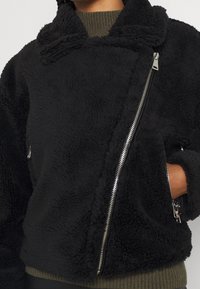 Black fleece jacket with a textured, plush surface. Features a shawl collar, diagonal zipper, and side pockets with silver hardware accents.