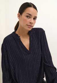 Blouse marine avec des rayures verticales argentées. Présente un col à volants, un décolleté en V et des manches amples. Texture de tissu douce.