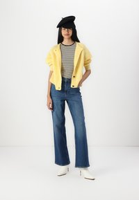 Cardigan en tricot jaune, t-shirt rayé noir et blanc, jeans évasés bleu, et bottes à talons blanches, accompagnés d'un béret noir.