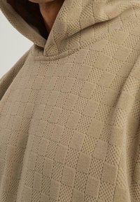Sweat à capuche beige en maille avec motif damier texturé, porté par une personne, montrant le haut du torse et le détail de la capuche.