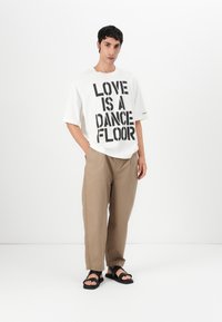 Balta didelė t-shirt marškiniai su ryškiu juodu tekstu "MEILĖ YRA ŠOKIO AIKŠTELĖ", derinami su smėlio spalvos kelnėmis ir juodais šlepetėmis. Paprastas fonas.